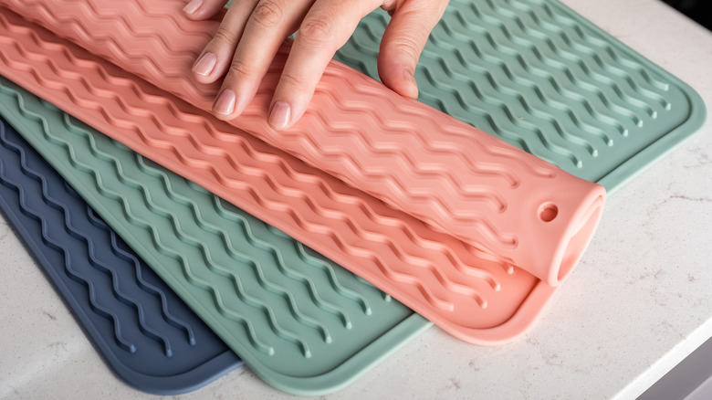 Three silicone dish mats of different colors with one being rolled up by a hand