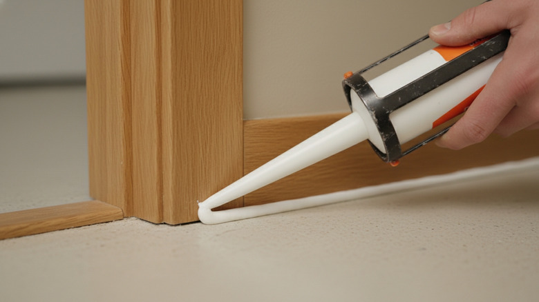 A hand holds and guides a caulk gun as it applies white caulk along brown baseboards