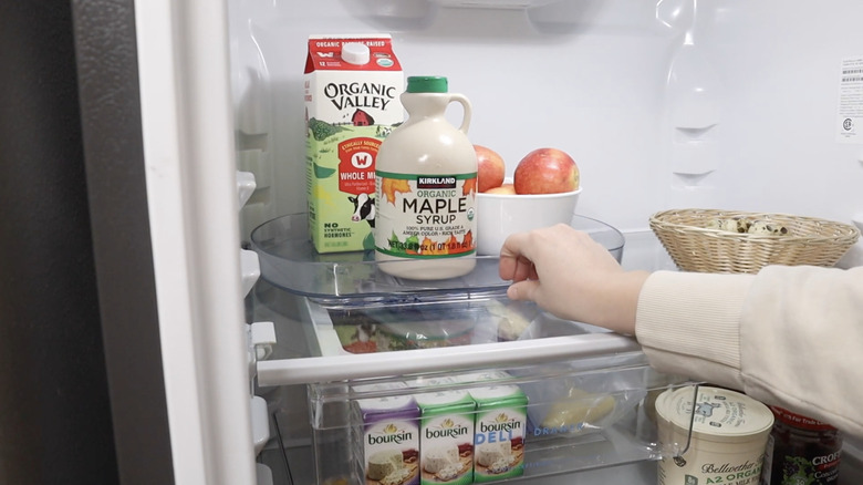 A lazy Susan organizer with mapple syrup, milk, and a bowl of apples sitting on it on a refrigerator shelf