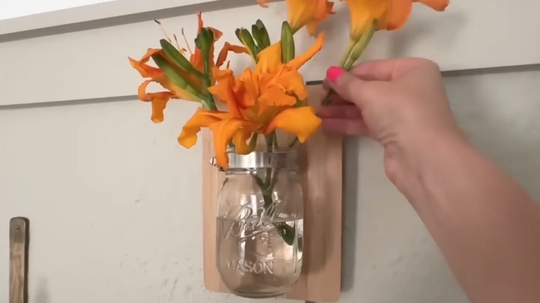Woman tucking orange flowers into Ball mason jar mounted on wood plaque on wall