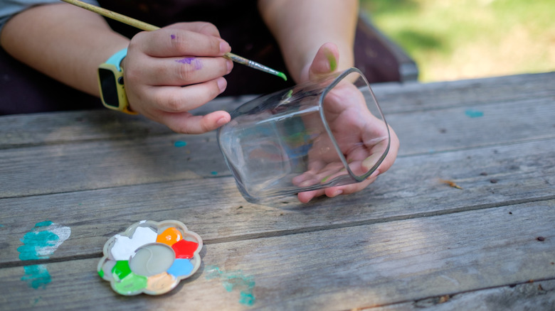 Person using a paint brush to apply paint on a square glass jar with a paint palate on a table in the foeground