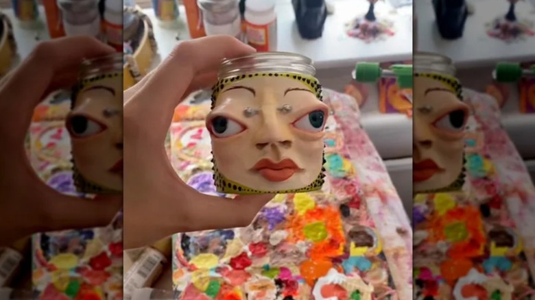Hand holding mason jar with eccentric clay art above colorful crafting table