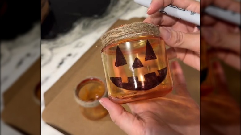 Woman designing orange-tinted jars with a marker-drawn Jack-o-Lantern face