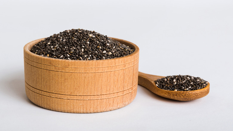 A wooden bowl filled with chia seeds next to a wooden spoon holding a scoop of chia seeds
