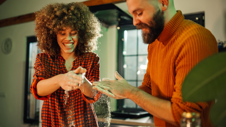 A couple in colorful outfits spread flour on a surface as they prepare to bake in a kitchen