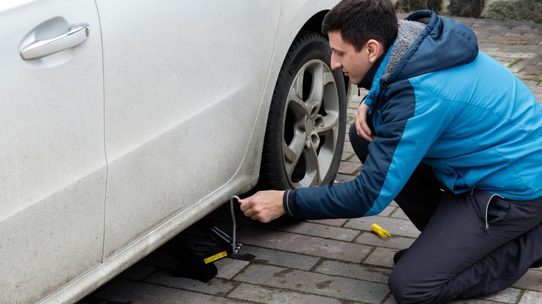A man works on his car in a driveway, with a stain beside the tire