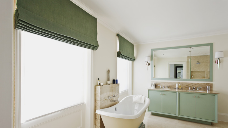A bathroom with a freestanding tub, sage green accents, and Roman blinds in the window