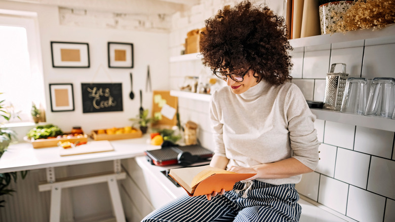 a woman sitting on a kitchen counter holding a cookbook