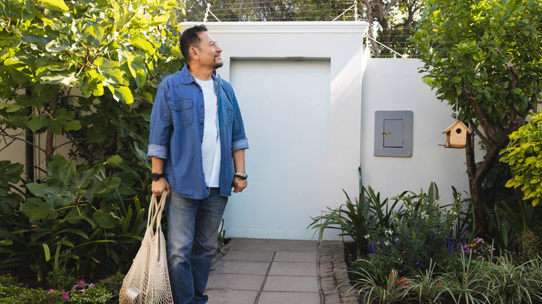 A man holds a shopping bag while standing on an old paver path in front of a small white building