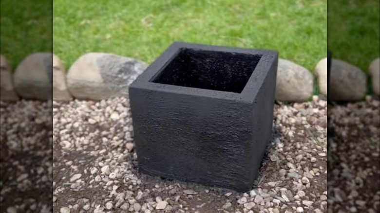 A black DIY planter made of concrete pavers sits in a bed filled with landscape gravel