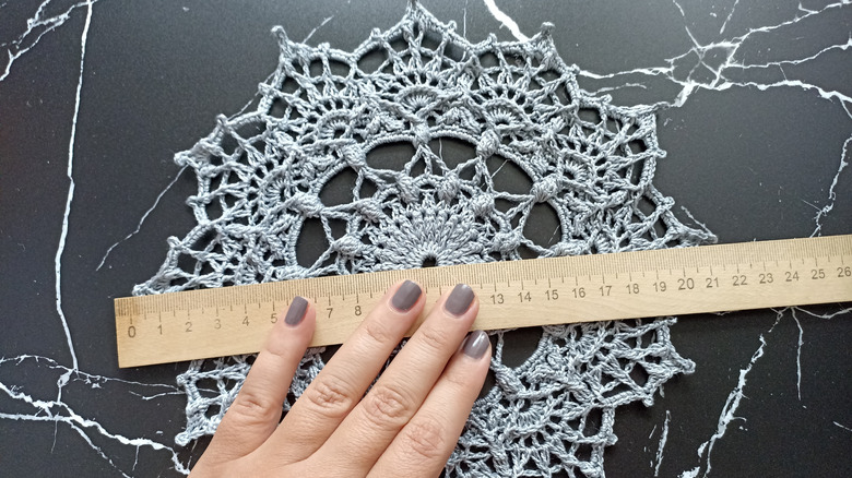 A hand with painted nails measures a lace doily on a table