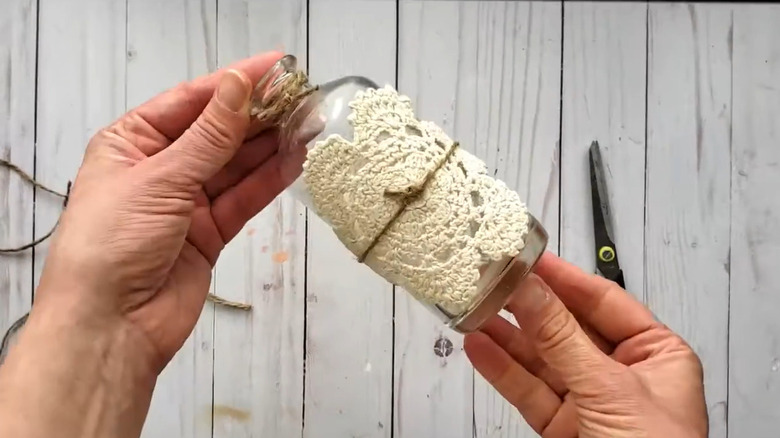 Man wrapping tiny jar in twine and lace doily over white wood background