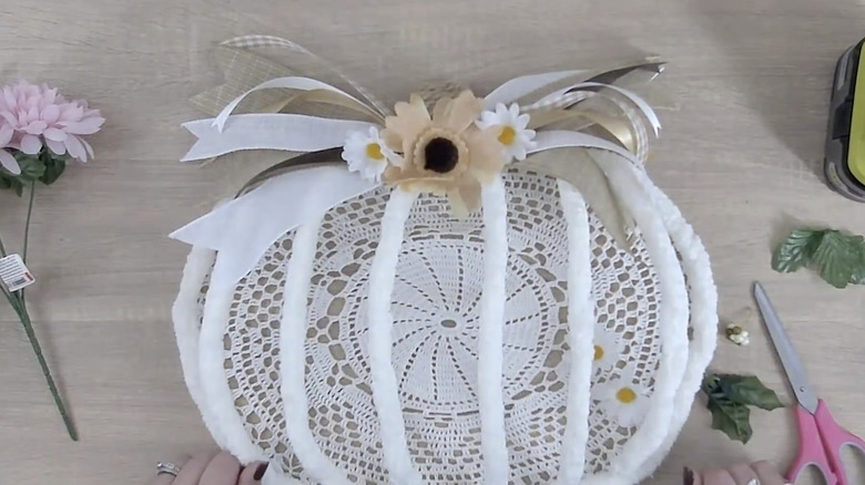 A 3d wire pumpkin frame accentuated with white yarn and fall accents, surrounded by a craft table with accessories and florals