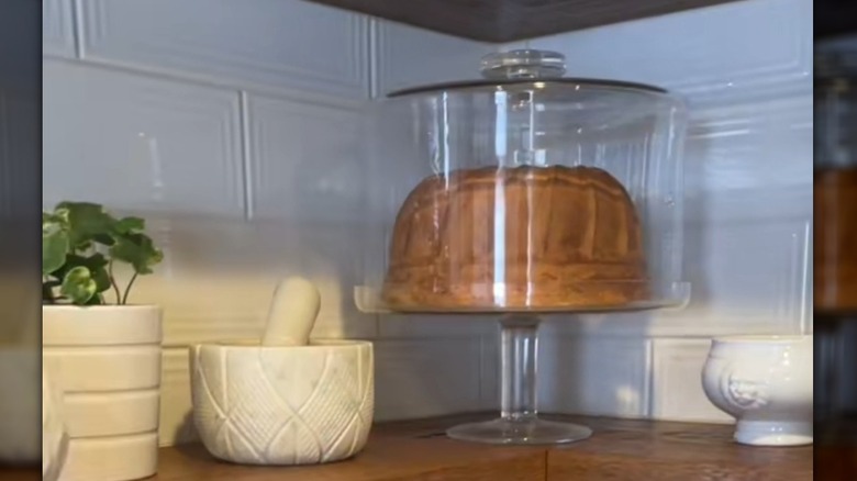 A real-looking imitation Bundt cake inside a glass cake stand on a kitchen shelf