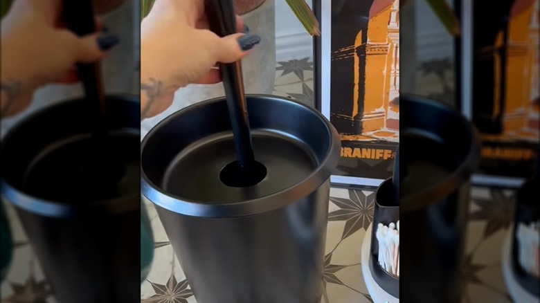 Creator holding the handle of a toilet plunger sitting inside a DIY holder made from a trash can