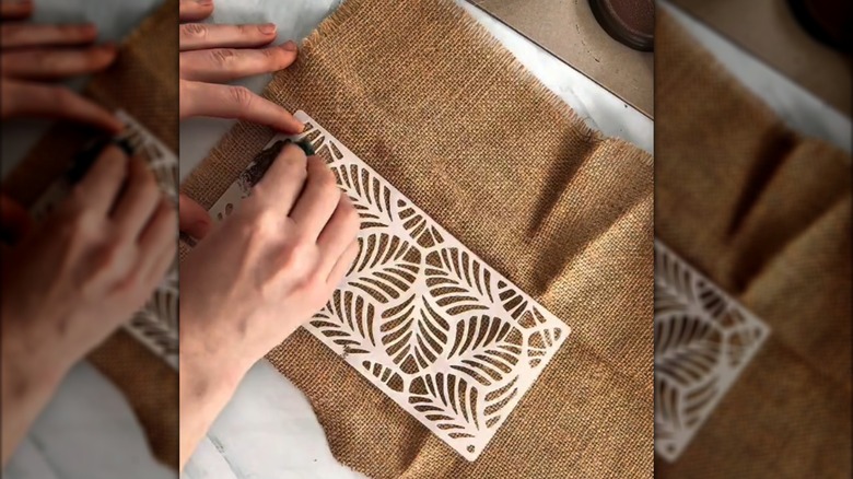 Woman's hands dabbing brown paint on burlap with stencil on white table surface