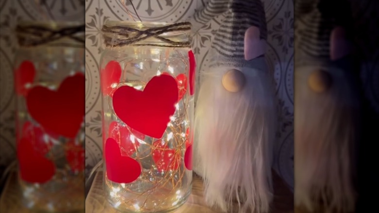 A glass jar with heart vinyl stickers on it and fairy lights inside beside a cute gnome