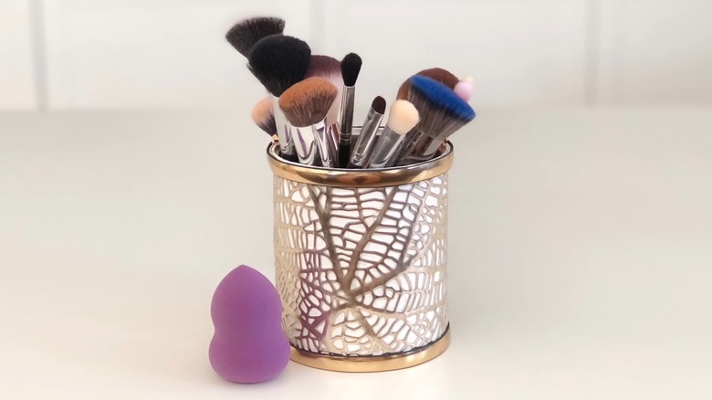 Makeup brushes inside a gold patterned holder with a beauty blender nearby