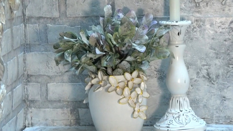Glass jar with pistachio shell flowers painted white and gold with a plant inside the jar, next to a candlestick