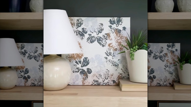 An abstract painted canvas sits on a wooden shelf behind a small lamp and potted plant