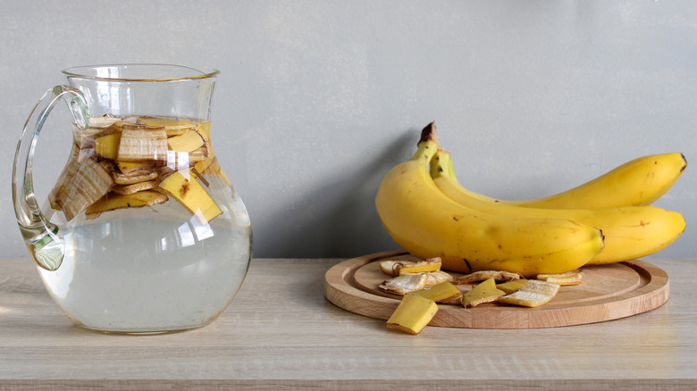 A jug of water and chopped banana peels next to bananas on a cutting board