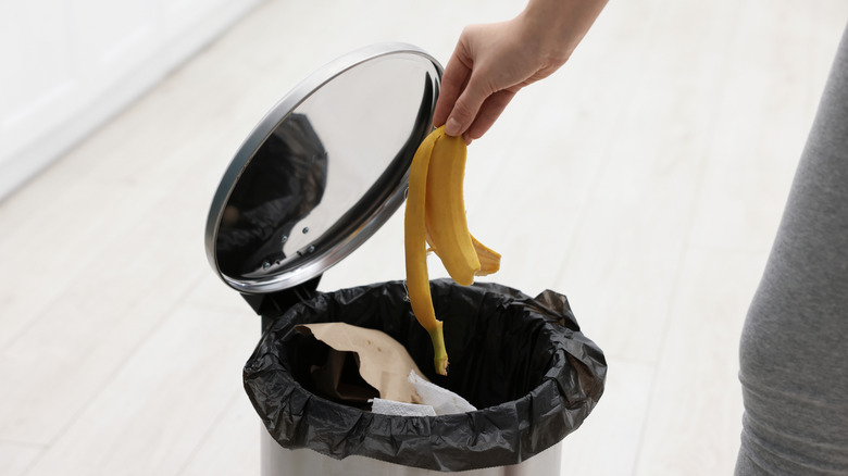 A person dropping a yellow banana peel into a trash can