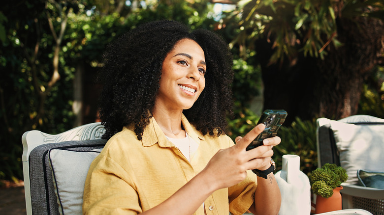 A woman smiles and thinks while looking at her phone in a lush backyard