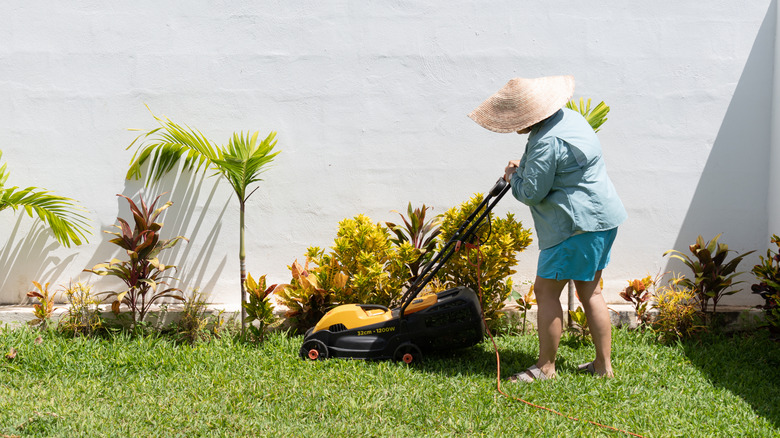 Older woman with sun hat pushing corded electric lawn mower beside houseplants