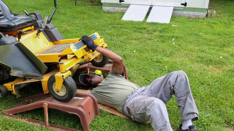 Man performing maintenance on riding lawn mower in lawn beside outdoor shed