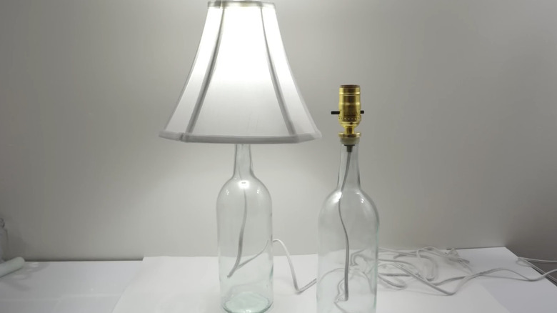 A clear wine bottle table lamp with a white shade next to a half-finished bottle lamp on a white surface.