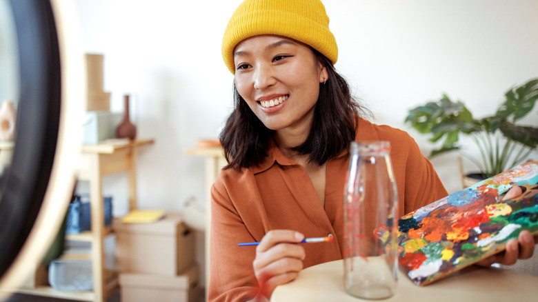 A person wearing a yellow hat paints an empty glass bottle using a colorful paint palette.