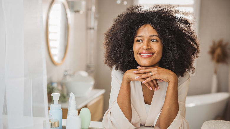 A woman smiles in her bathroom, deep in thought, with a selection of toiletries on the countertop