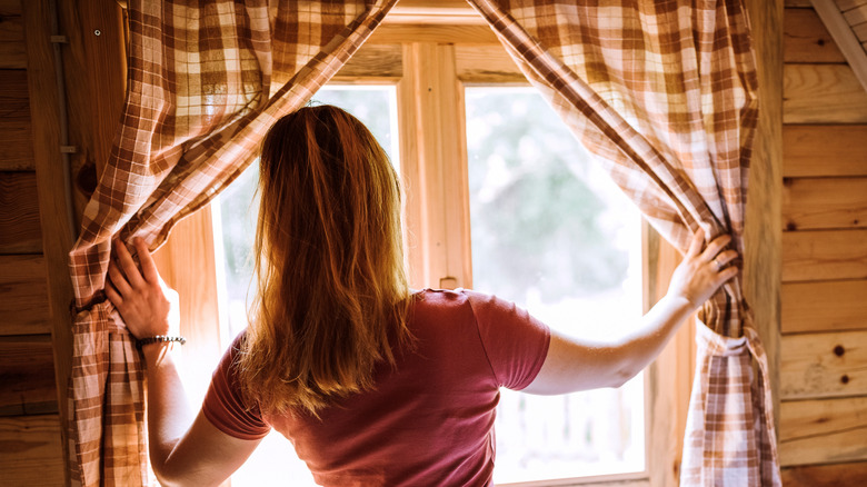 A woman draws back plaid curtains and looks out the window.