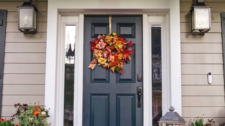 A fall wreath on a blue door surrounded by autumnal plants and two large front lights and black shutters