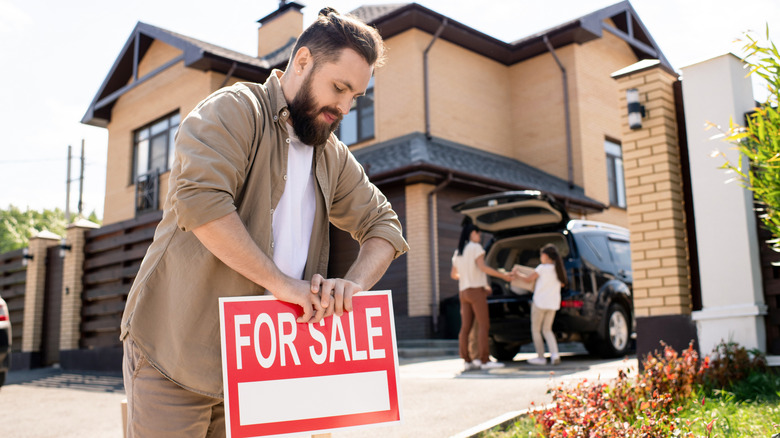 A man places a "For Sale" sign in front of a brown home