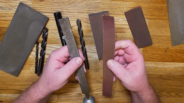 Man sharpening drill bit with aluminum oxide abrasive paper in workshop setting.