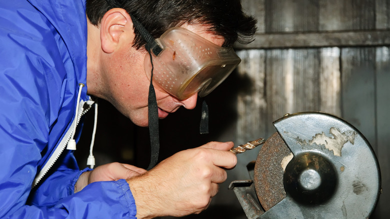 Man with goggles, sharpening a rusty drill bit using a bench grinder.