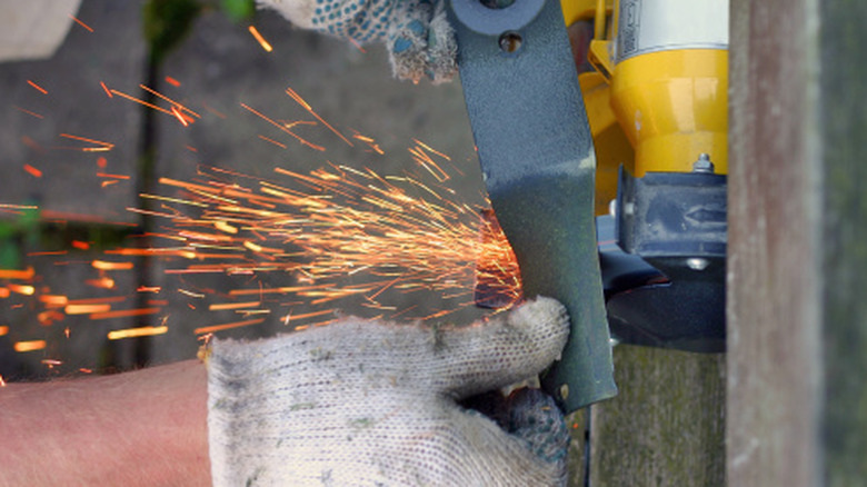 Hands sharpening a lawn mower blade with a bench grinder, with sparks flying off