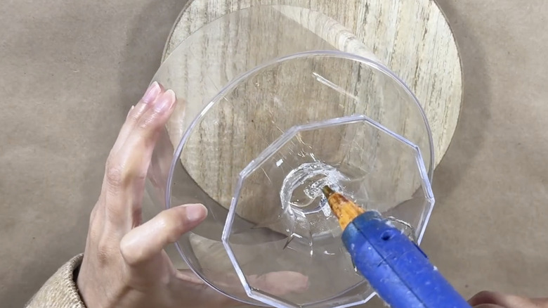A woman's hand with a hot glue gun applying adhesive on a plastic container and circular wooden tray