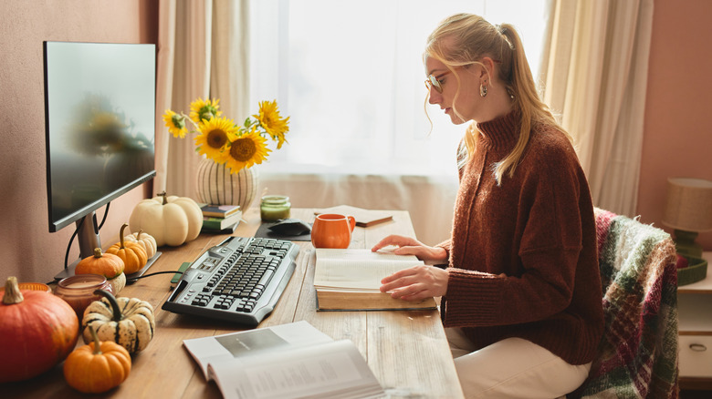 A woman sits by her fall-themed desk, with pumpkins, a mug, candles, and sunflowers resting on it