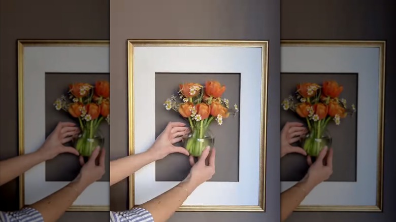 A woman's hand placing a small floral vase within a gold picture frame fastened to the wall