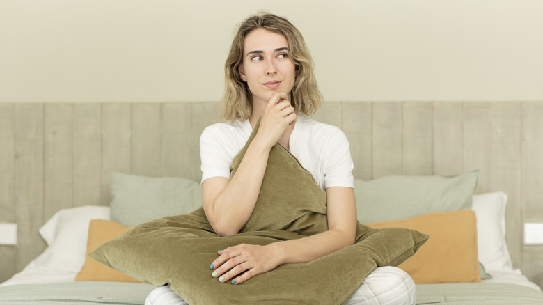 A woman sits on a bed, hand on chin, and thinking