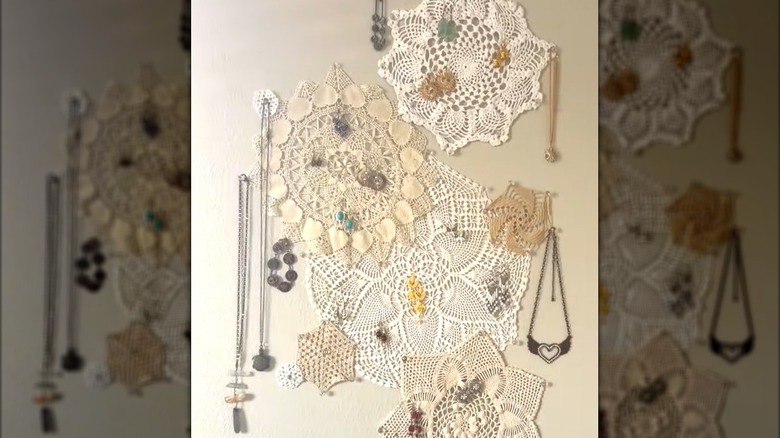 Assorted lace doilies pinned to beige wall with hanging necklaces, earrings, and bracelets