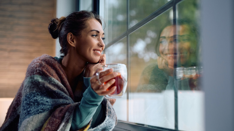 Young smiling woman drinking tea and looking through the window at home.