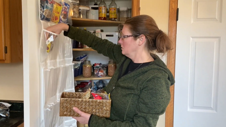 A woman puts snacks in her plastic shoe rack hanging on her pantry door, organizing snacks.