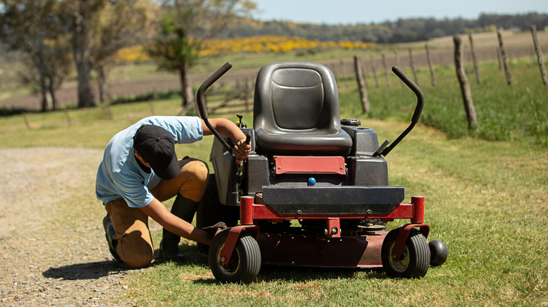 Man wearing a hat checking the blades of a sit on lawn mower