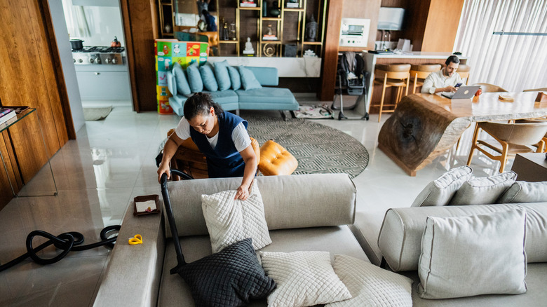 A housekeeper vacuums a sofa while a homeowner tries to work on a laptop sitting on a nearby table.