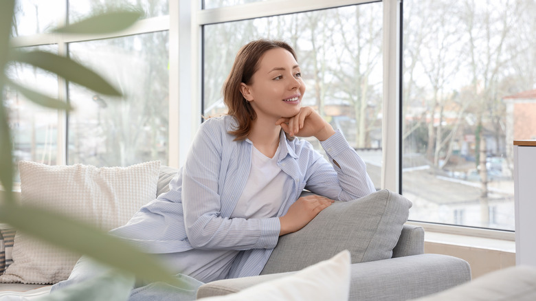 A person sitting on a gray couch looking out windows with a plant in the foreground