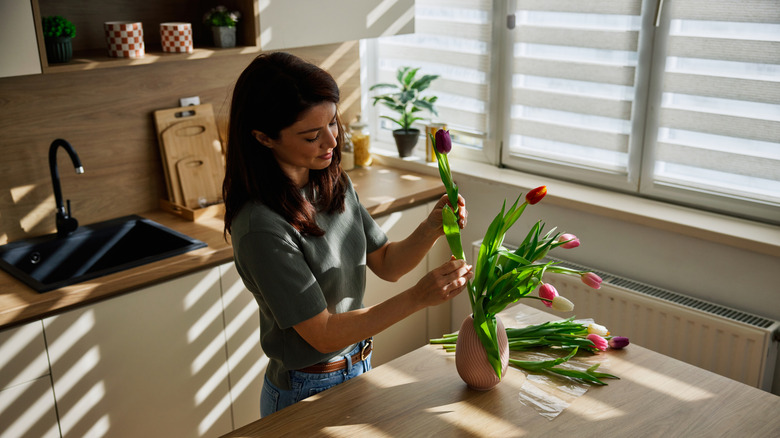 A woman adjusting flowers in a kitchen with window blinds providing shade.