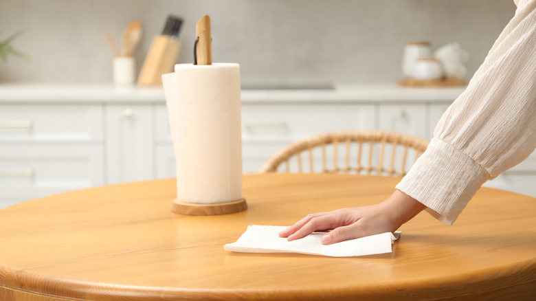 Wooden paper towel holder on a table and a woman wiping the table with a paper towel